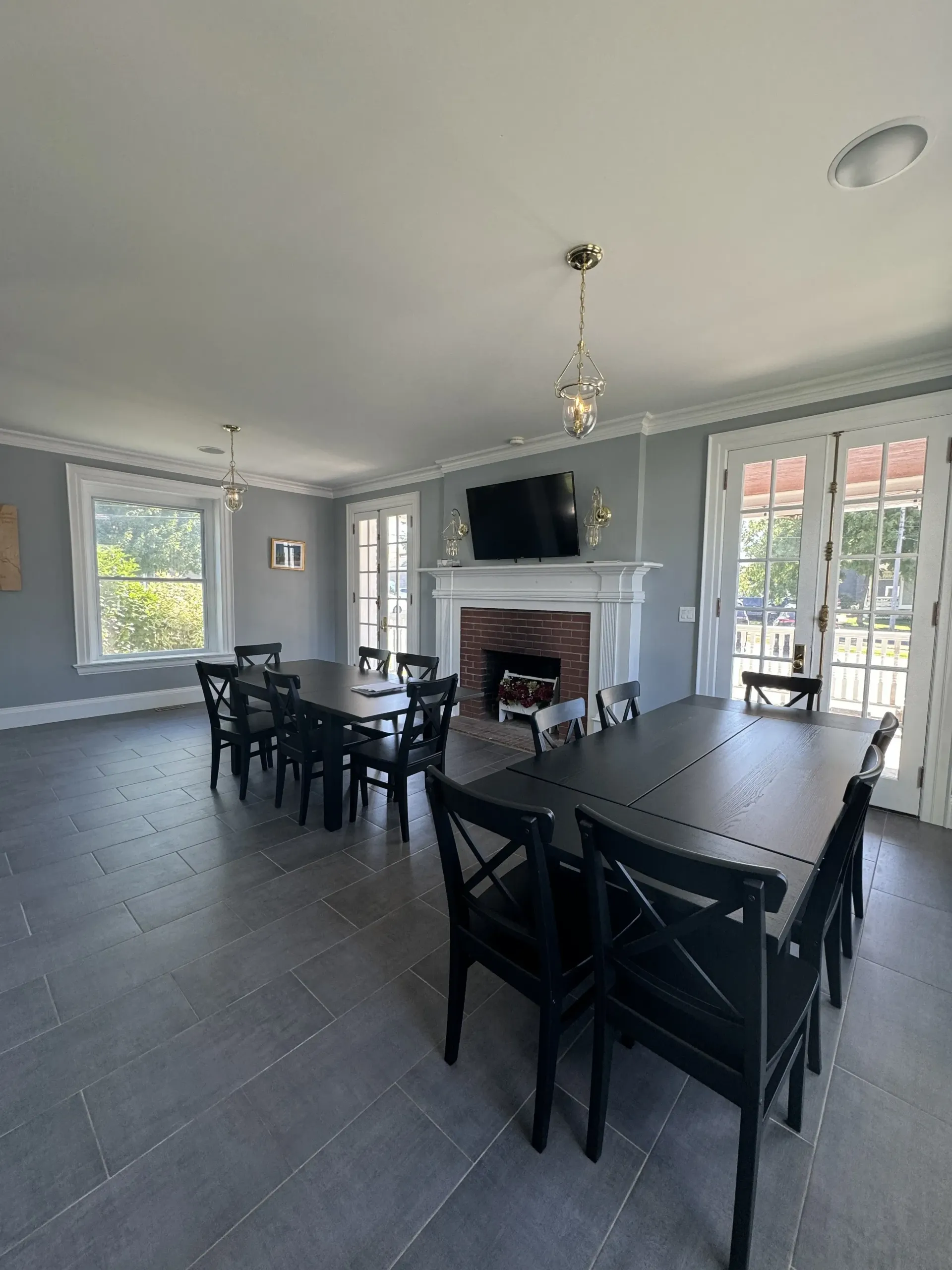 A bright dining room with gray walls, tile floors, a fireplace with a mounted TV, and two black wooden dining tables.