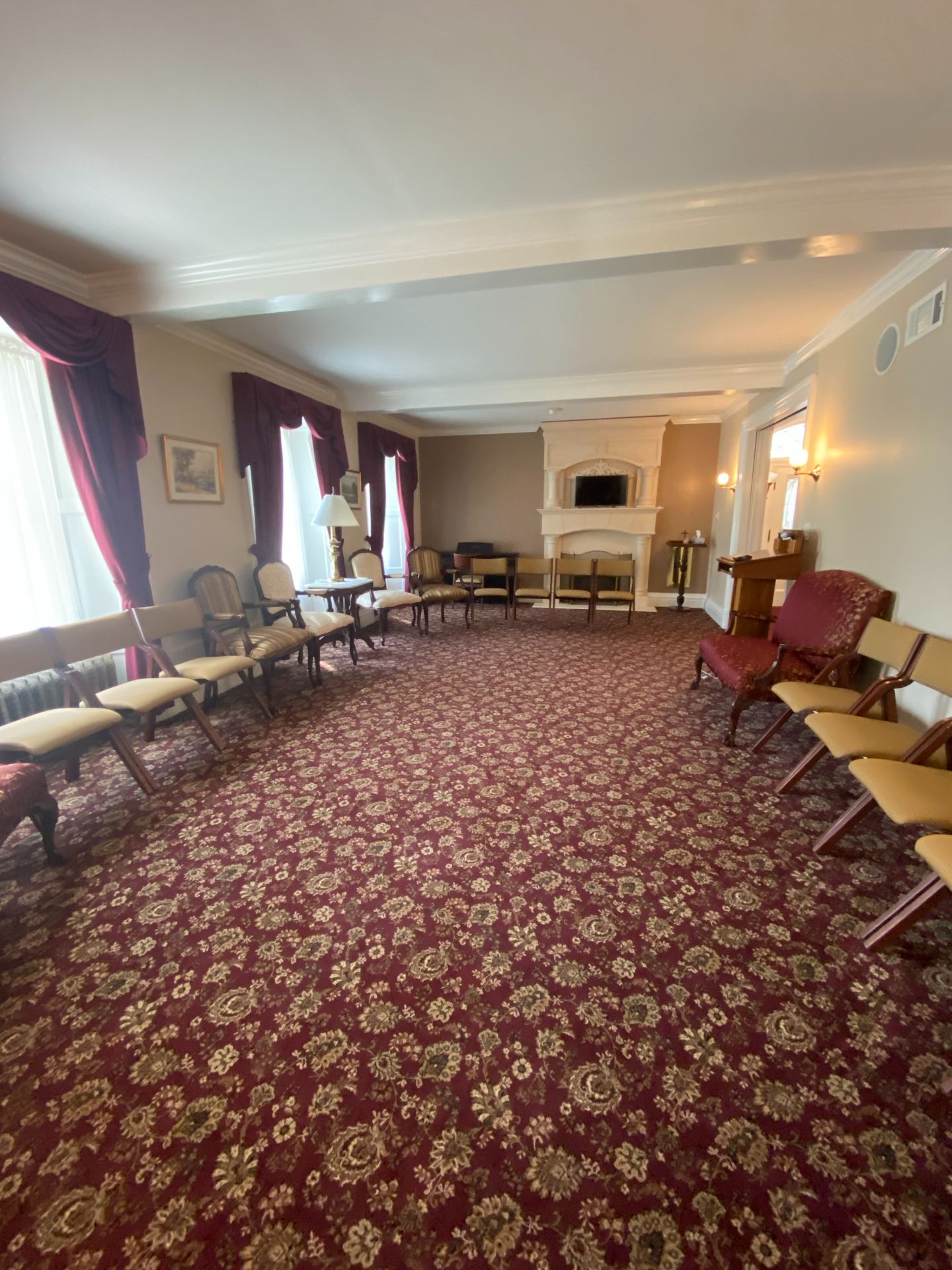 A spacious, carpeted room with maroon floral patterns, featuring rows of tan chairs, dark curtains, and a fireplace.