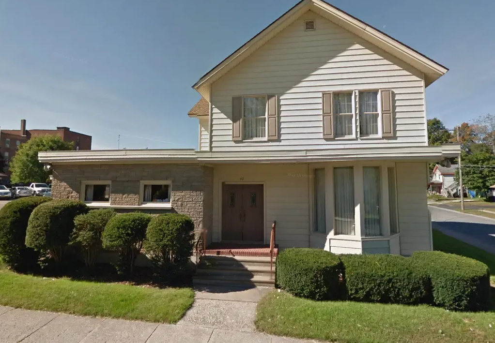 A two-story white house with a flat-roofed stone wing and a front porch, set back from a sidewalk with manicured bushes.