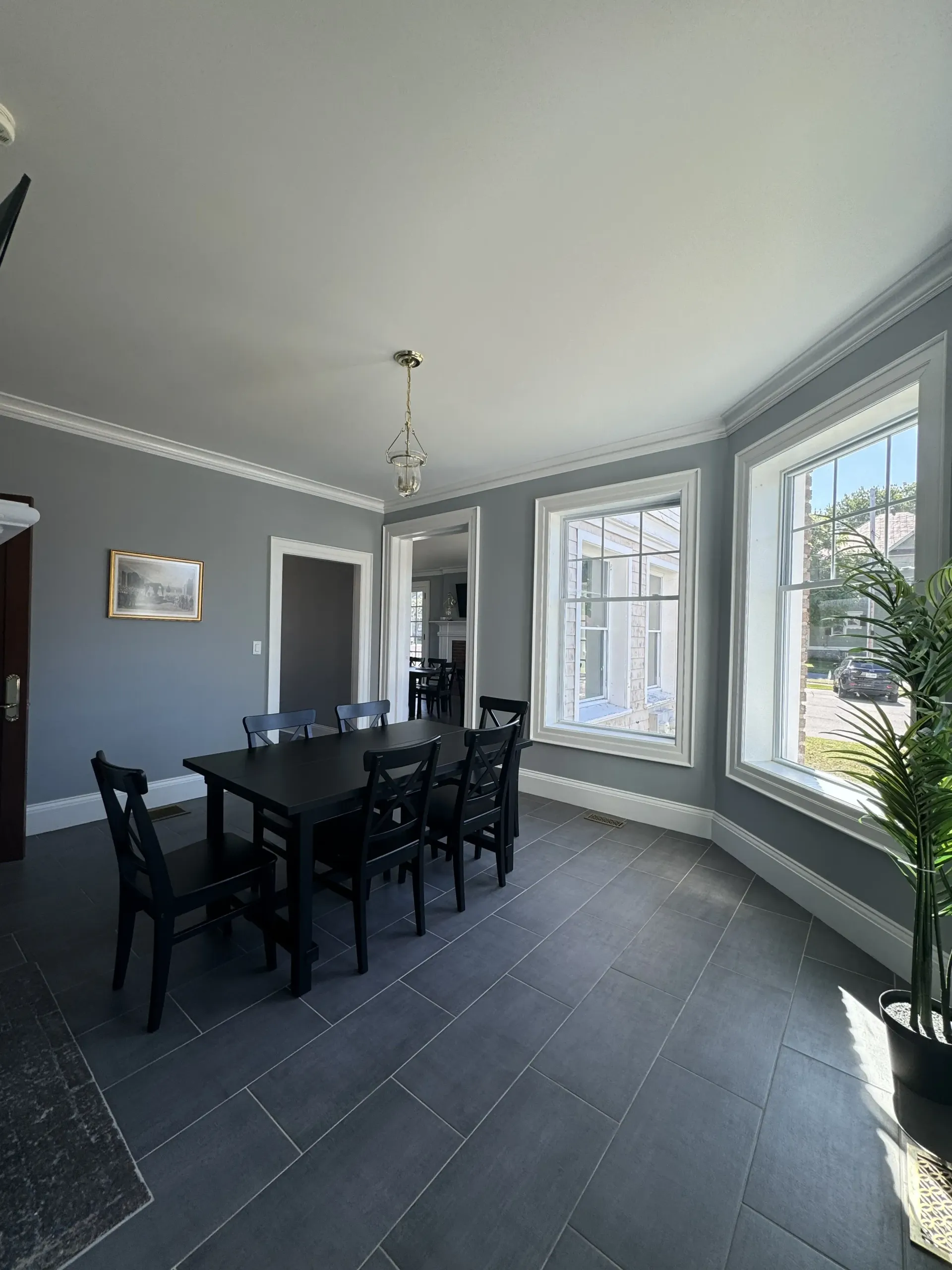 A dining room with gray walls, dark tiled floors, a black dining table and chairs, and bright windows on the right.