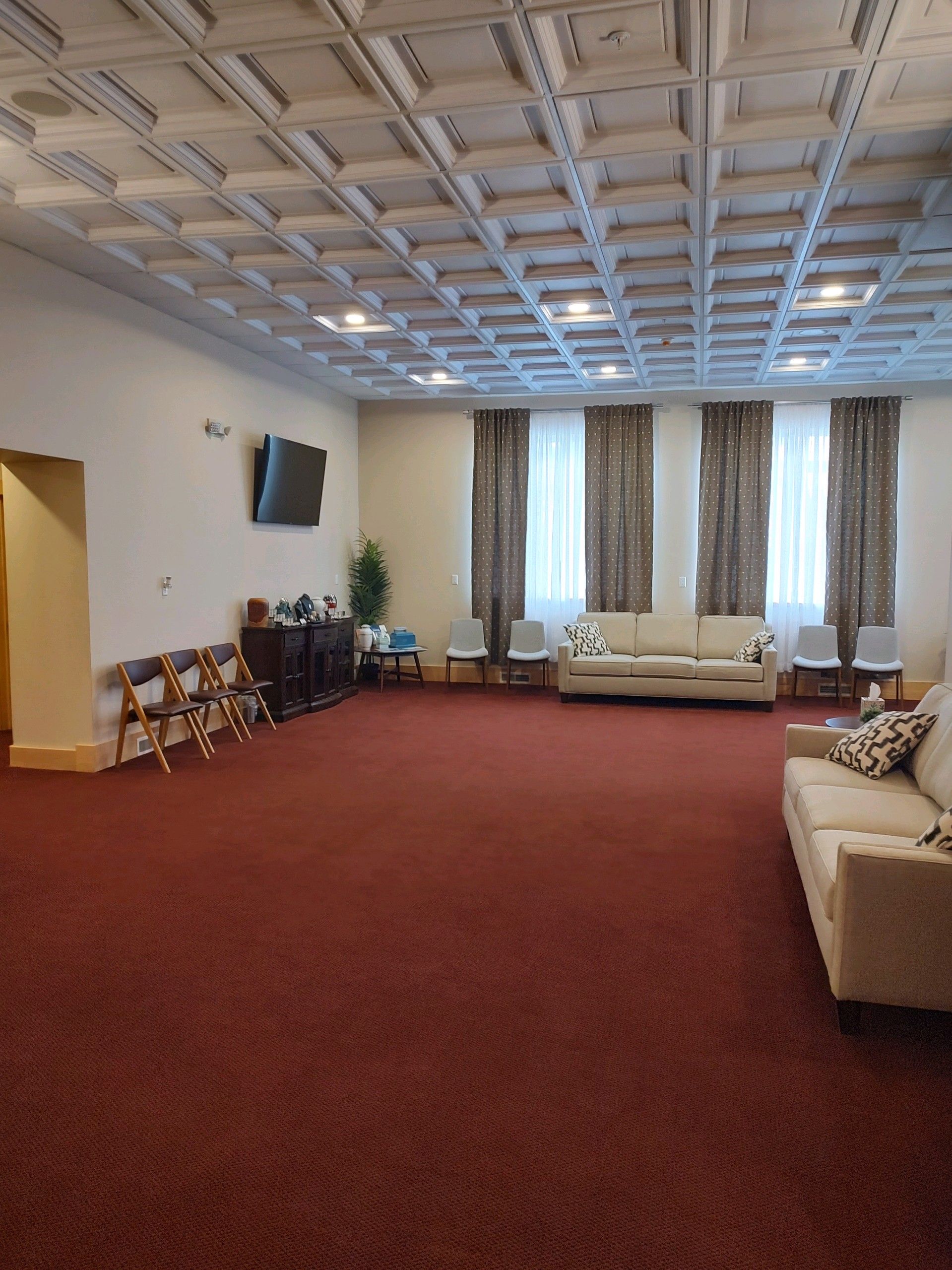 A large, sparsely furnished waiting area with red carpeting, a coffered ceiling, two beige couches, and chairs.