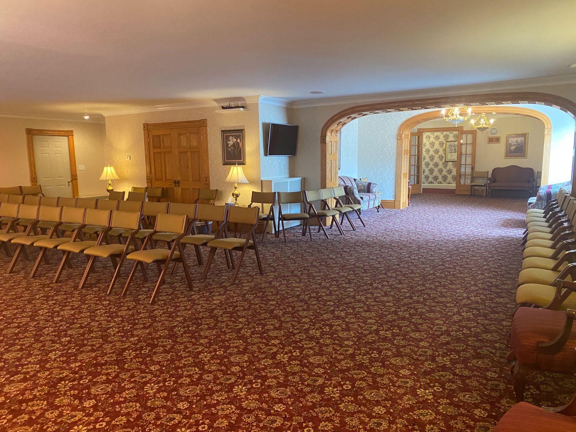 A large event room with rows of chairs on red patterned carpet, leading to a wooden arched opening into another space.