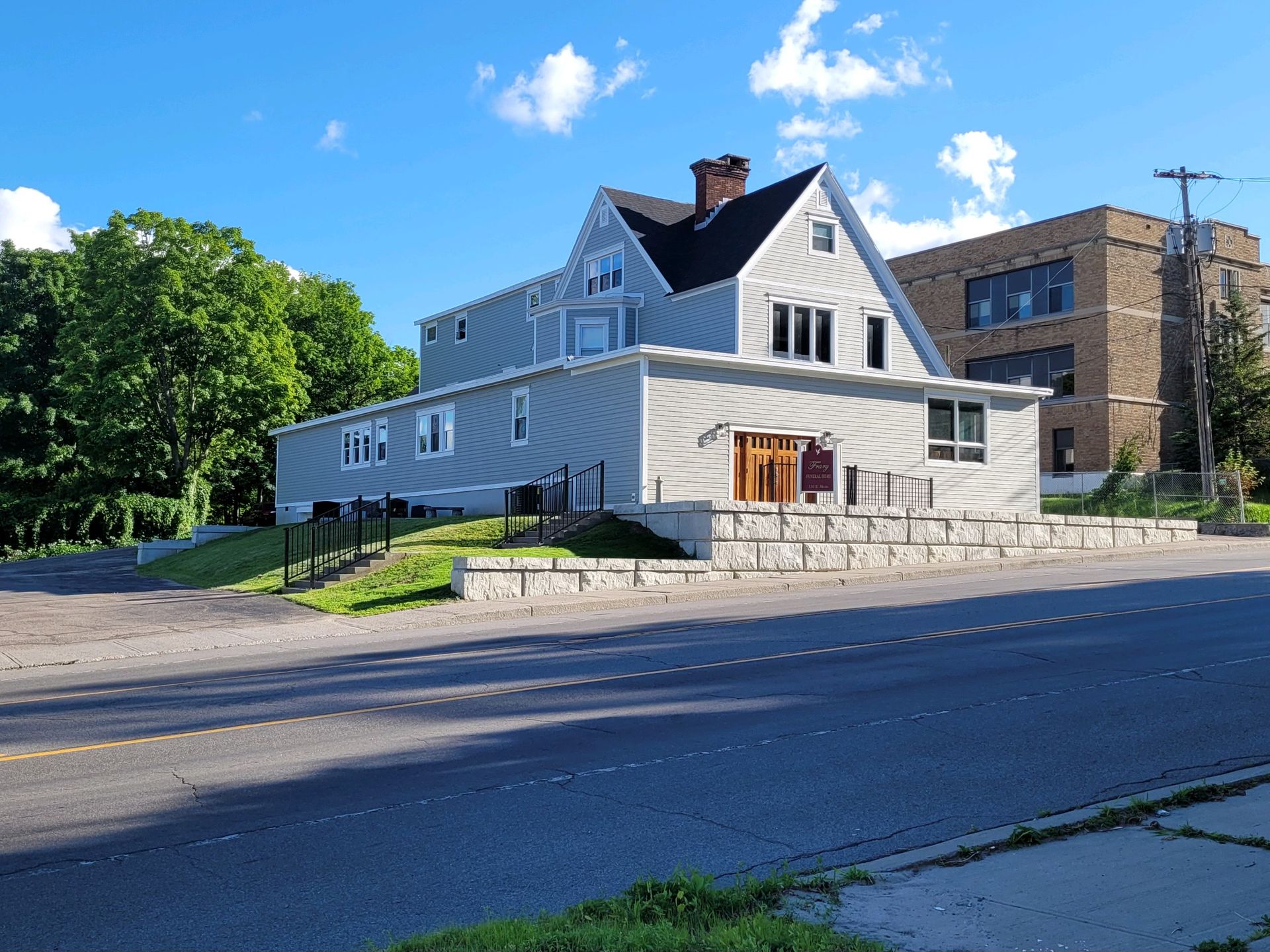 A light blue house with a dark roof sits on a stone foundation next to a brick building along a sunny street.