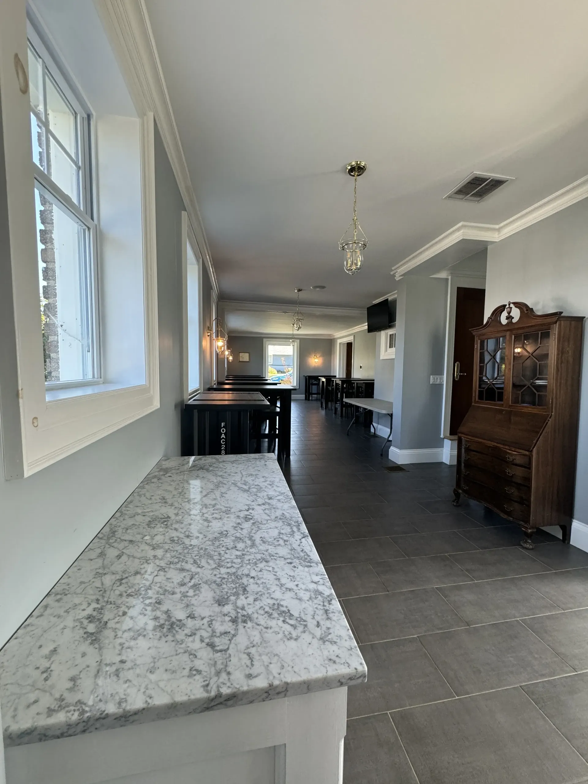 A light-filled hallway with a marble-topped counter, large windows, dark tiled floors, and a wooden hutch.