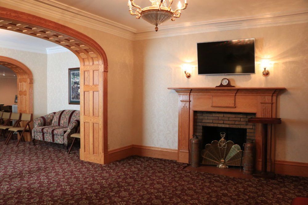 A wood-paneled fireplace and TV under a chandelier in a room with a patterned carpet and an arched doorway to a lounge.