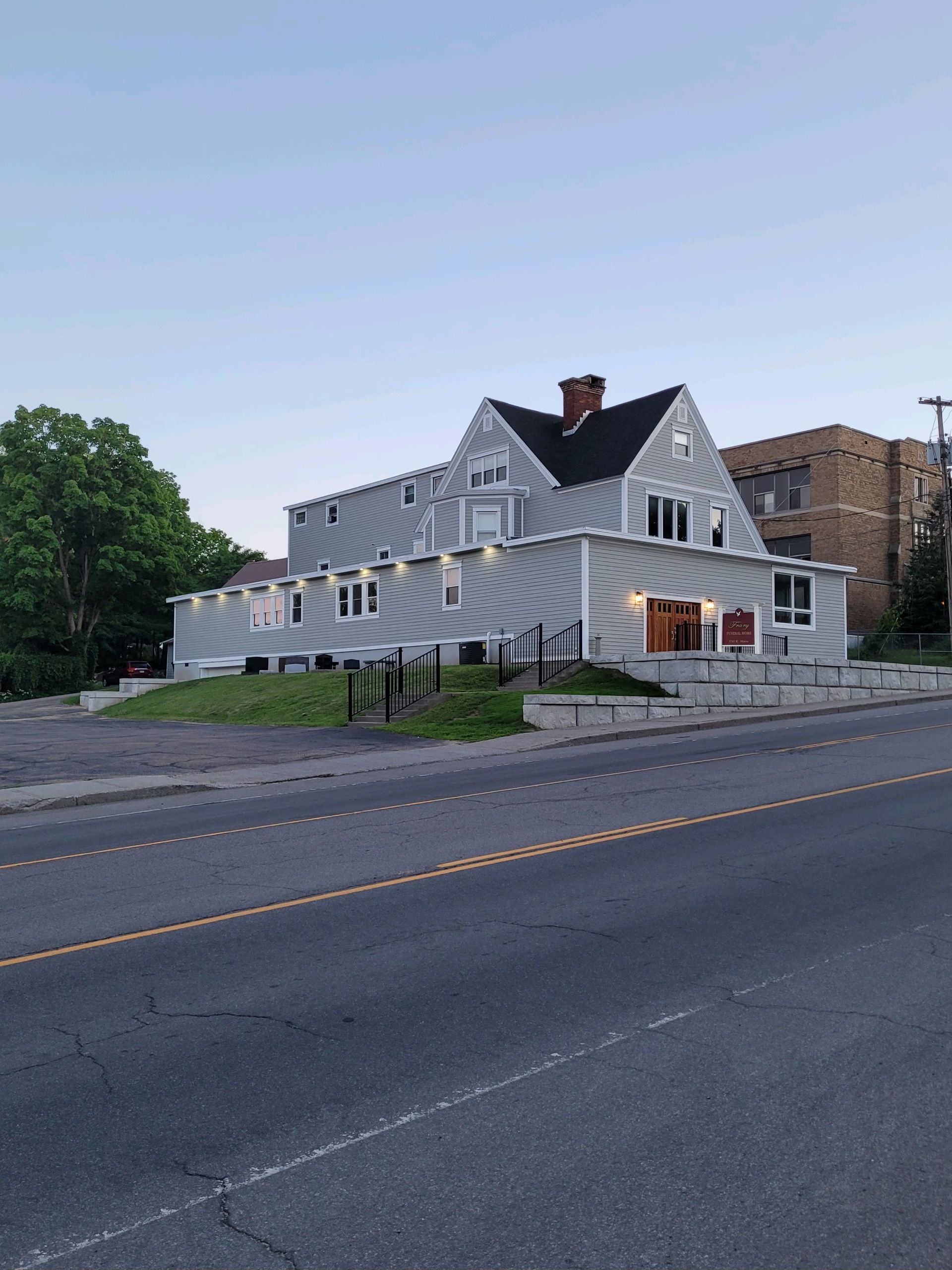 A light gray, two-story house with a black roof sits along a road at dusk, with exterior lights on.