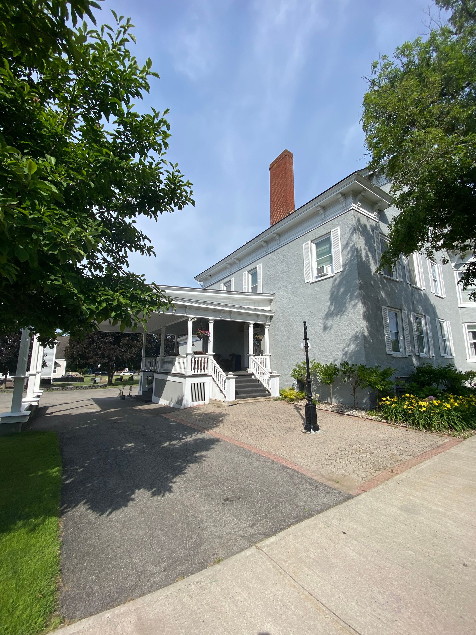A gray, historic-style two-story house with a white front porch, stone chimney, and a gravel driveway on a sunny day.