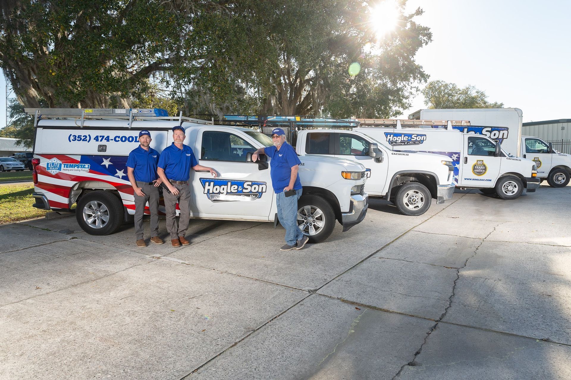Three men in blue shirts stand by service trucks with company logo. Sunlit parking lot setting.
