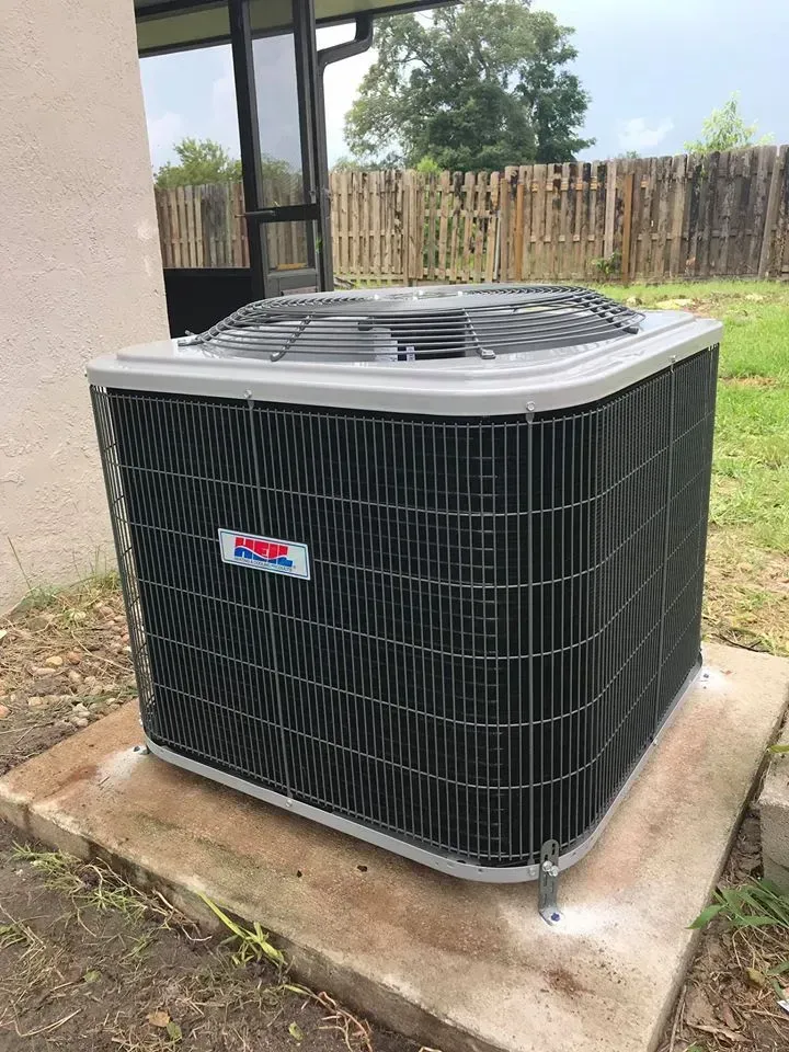 An air conditioning unit outdoors on a concrete pad, with a fence and building in the background.