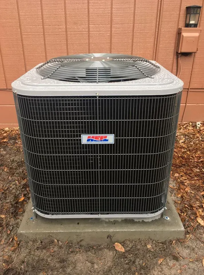 Air conditioner unit, gray and black, on a concrete pad, against a brown wall.