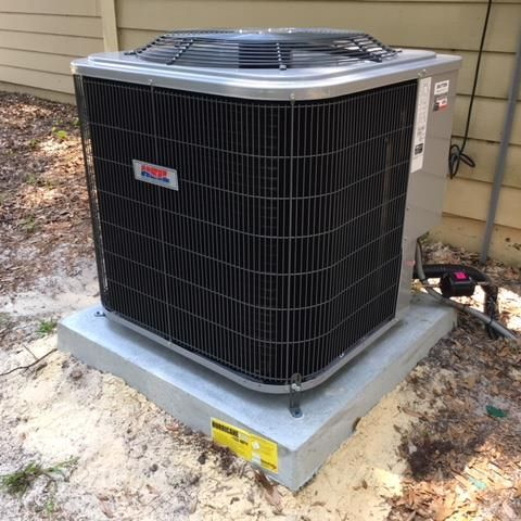 Exterior air conditioning unit on a concrete pad, surrounded by sand and foliage.