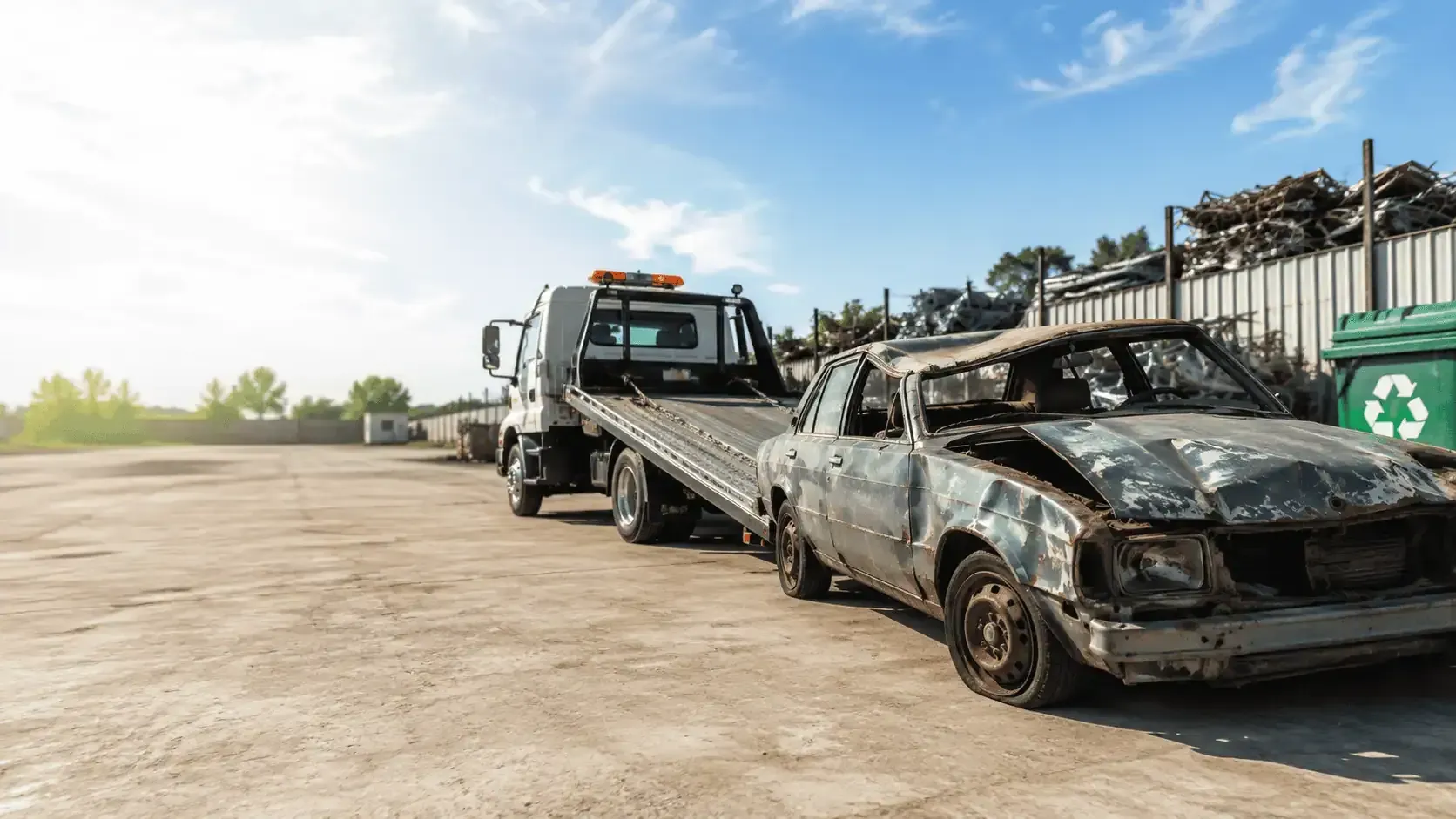 Tow truck loading a burned-out car at a scrapyard under a bright sky