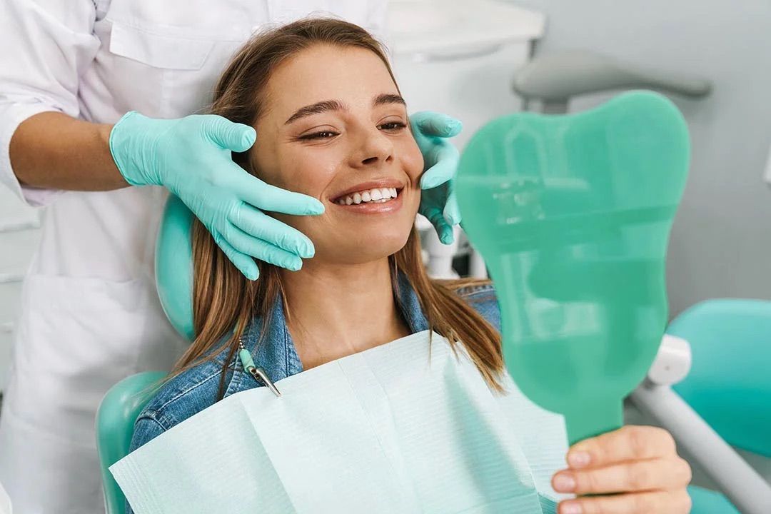 A woman is sitting in a dental chair looking at her teeth in a mirror.