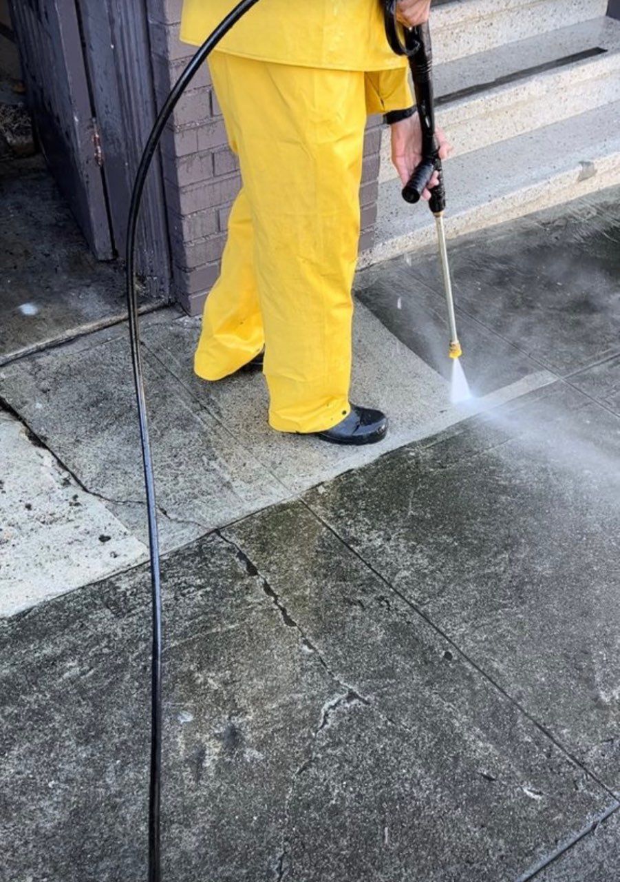 A person in yellow pants is using a high pressure washer to clean a sidewalk.
