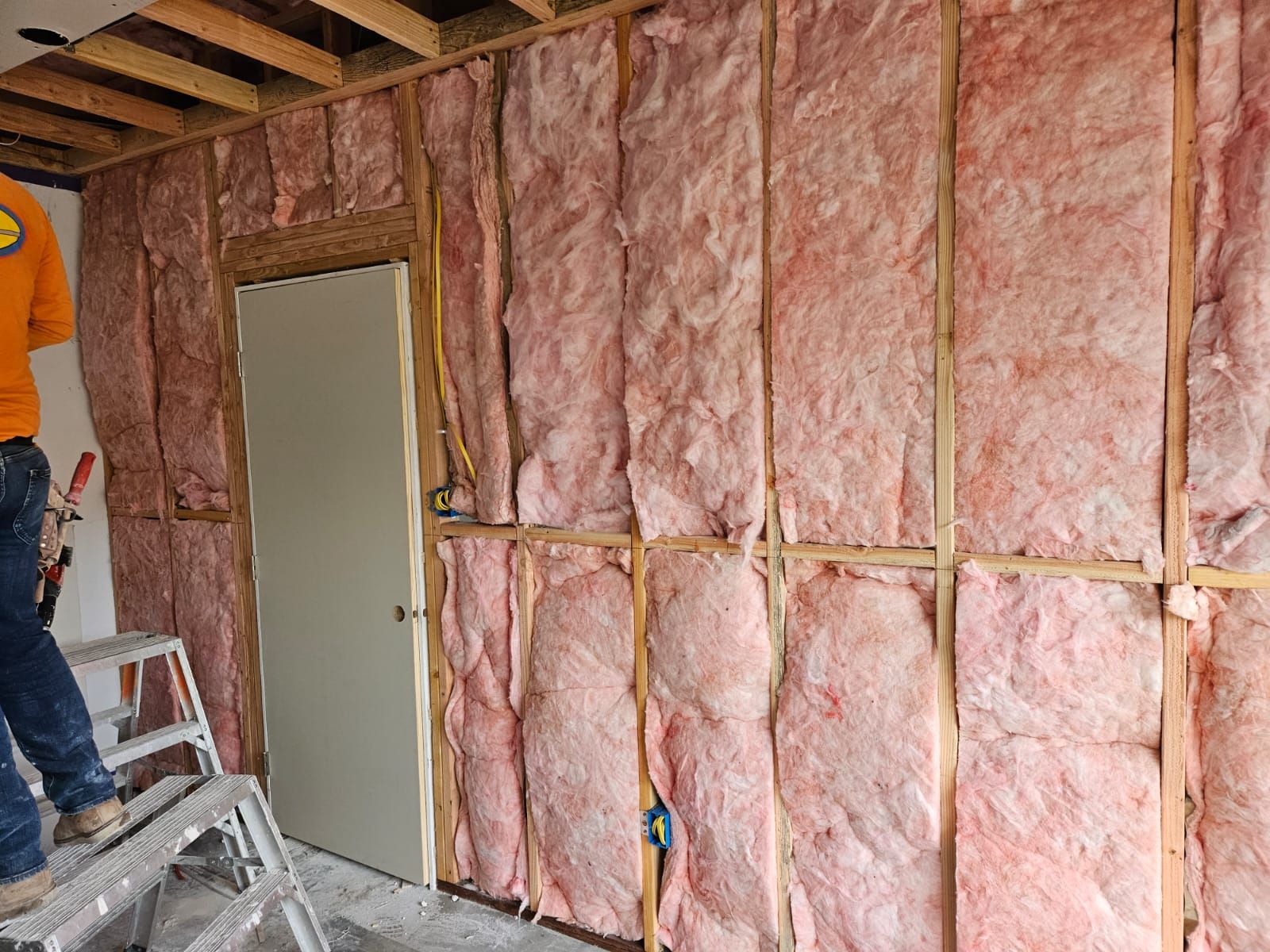 A man is standing on a ladder in front of a wall covered in pink insulation.