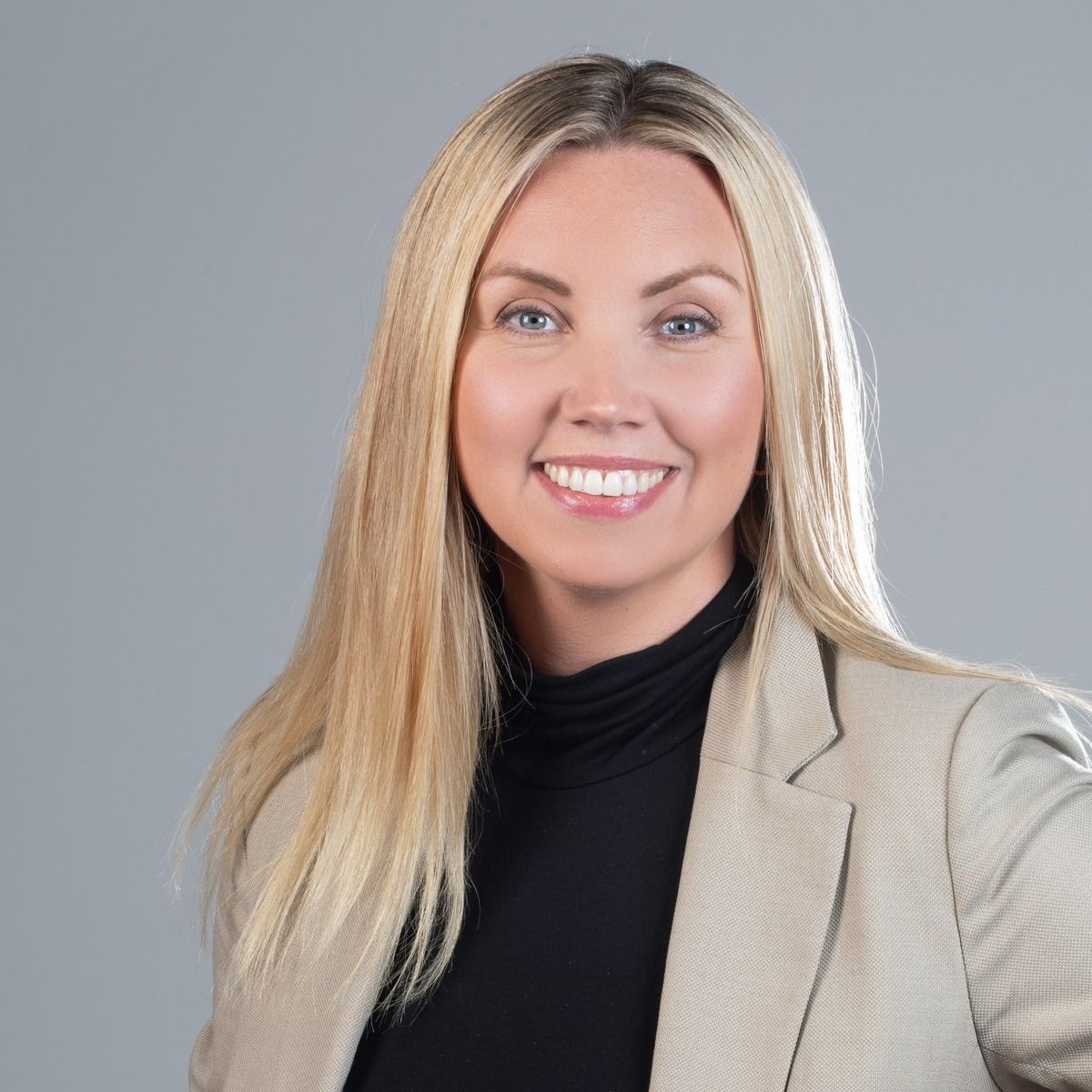 Woman with blonde hair smiles, wearing a black turtleneck and tan blazer against a gray background.