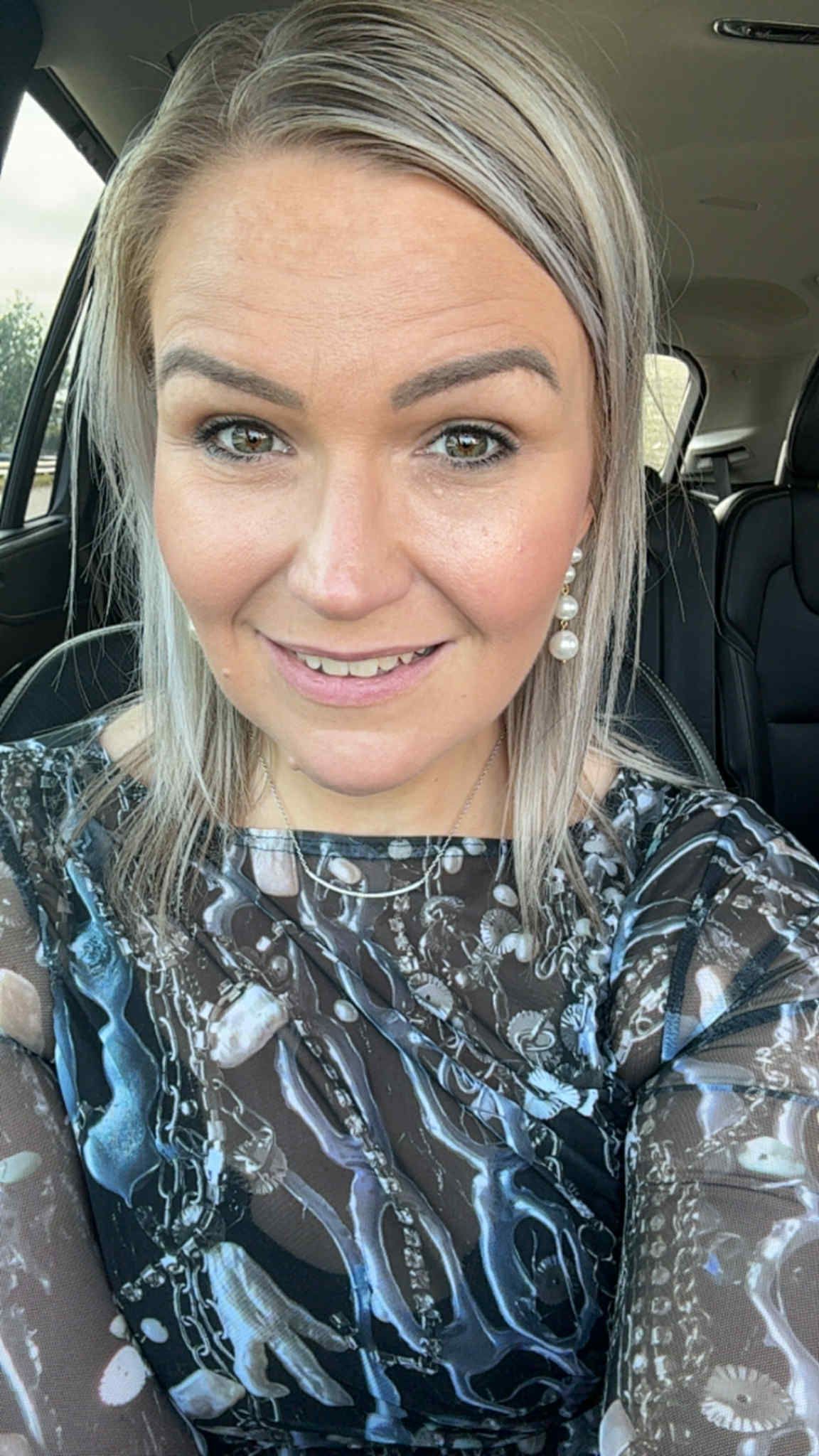Woman in a car smiling, with blonde hair, wearing earrings and a patterned top.