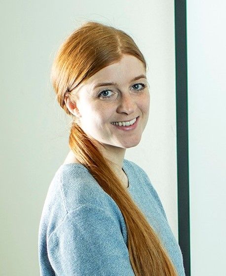 Woman with red hair in a ponytail smiles, wearing a blue sweater, in front of a white wall.