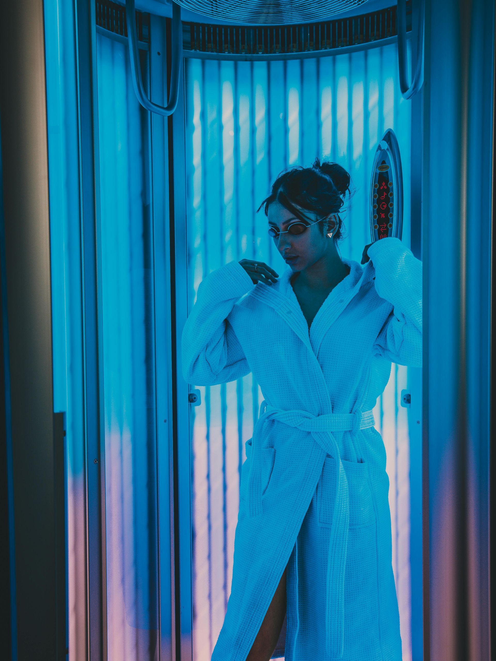 Woman in a bathrobe in a tanning bed, lit with blue lights.