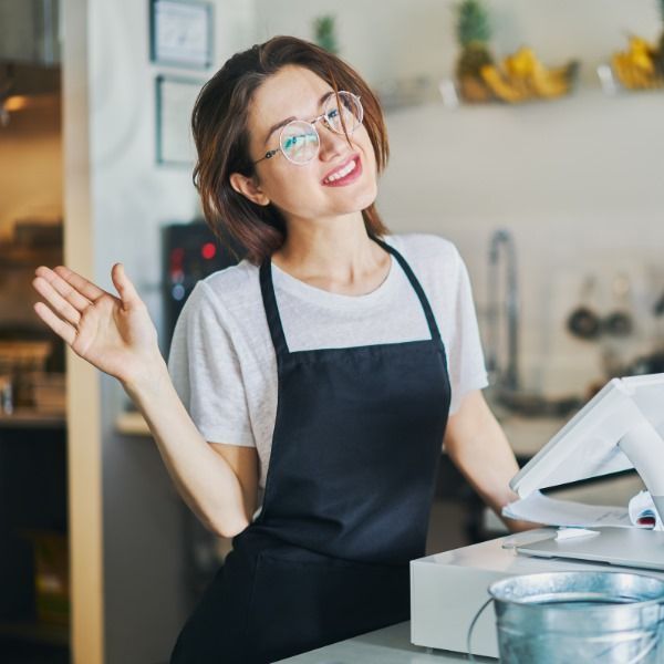 A woman wearing glasses and an apron is standing in front of a cash register.