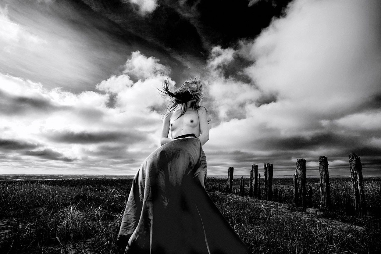 Een vrouw in een lange rok, met ontbloot bovenlijf, staat in een veld met houten palen onder een bewolkte hemel.