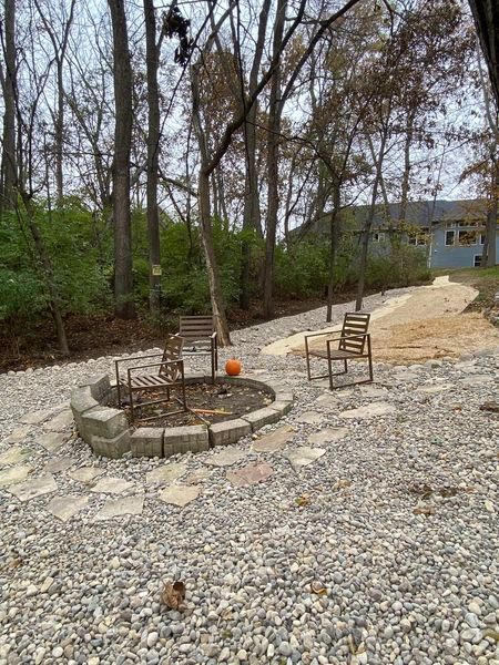 A fire pit in the middle of a gravel yard with chairs and a pumpkin.