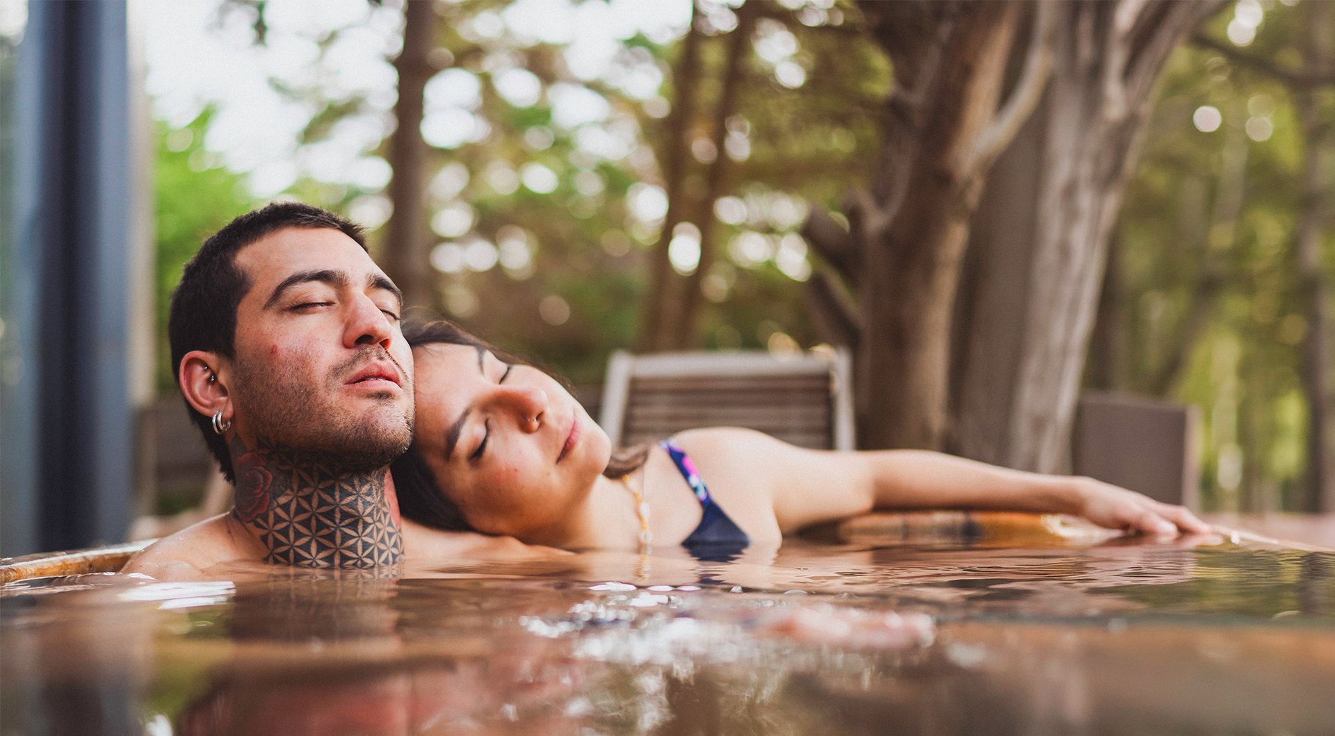 Una pareja con los ojos cerrados, relajándose juntos en un jacuzzi al aire libre rodeado de árboles.