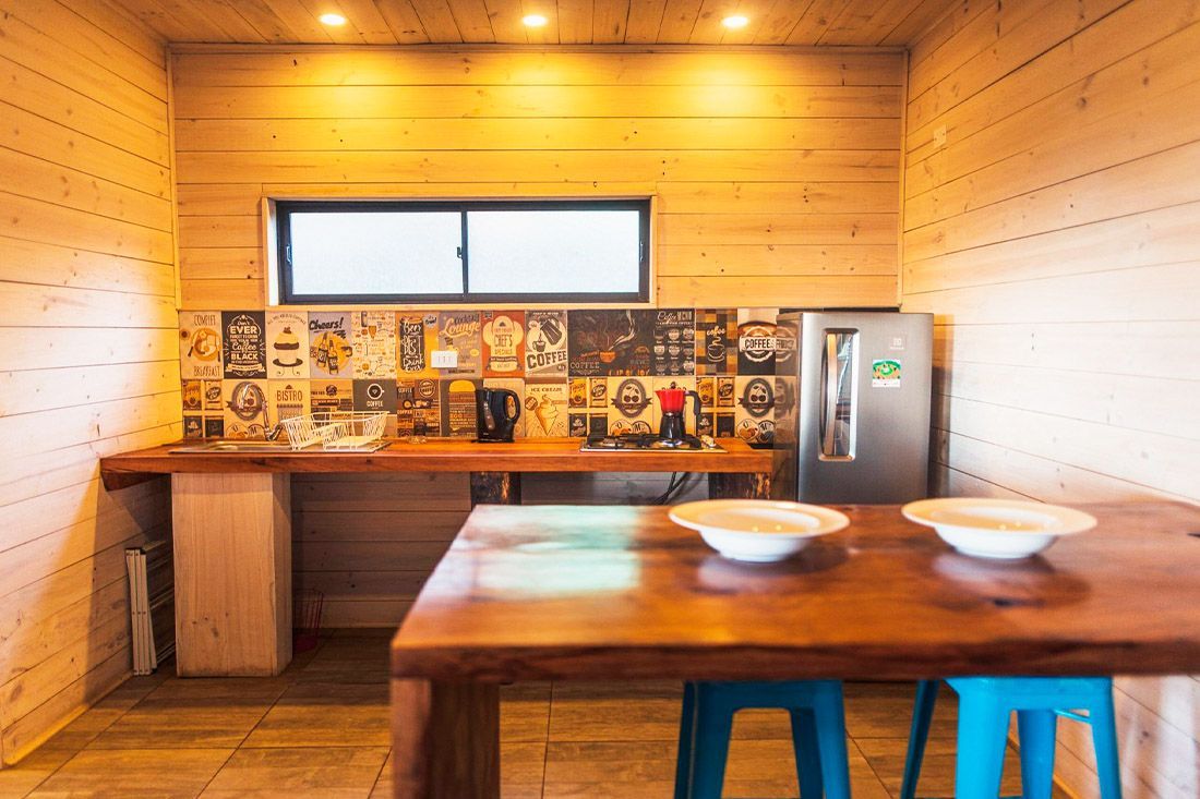 Una cocina rústica con paneles de madera, una mesa de madera, dos taburetes azules, un refrigerador plateado y un protector contra salpicaduras estampado.