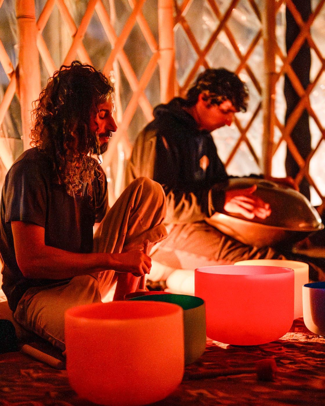 Dos personas tocan instrumentos musicales, incluido un handpan y varios cuencos cantores brillantes y coloridos en una yurta.