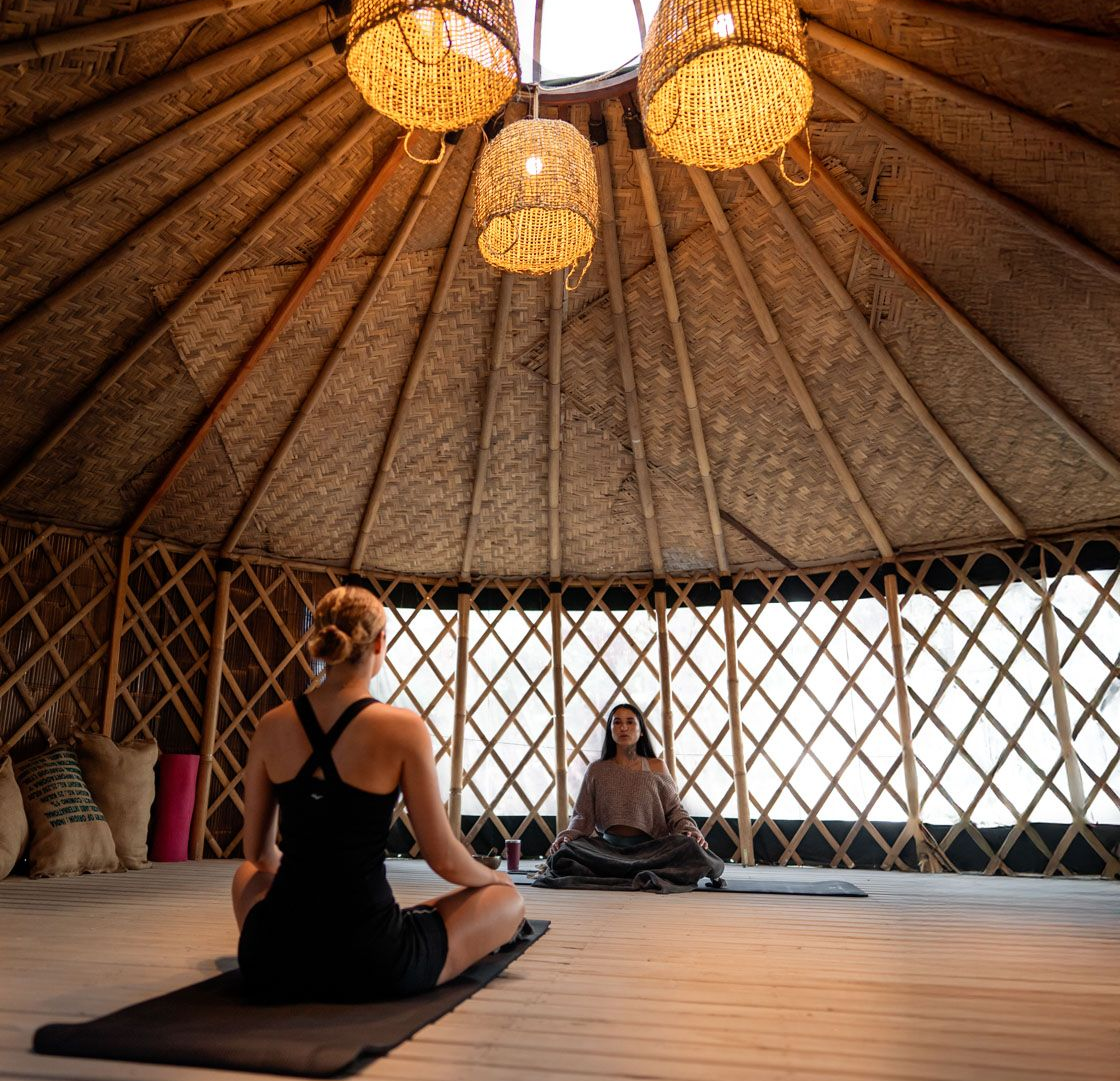 Dos personas se sientan sobre colchonetas de yoga en una yurta circular de madera iluminada por tres cálidas luces colgantes tejidas.