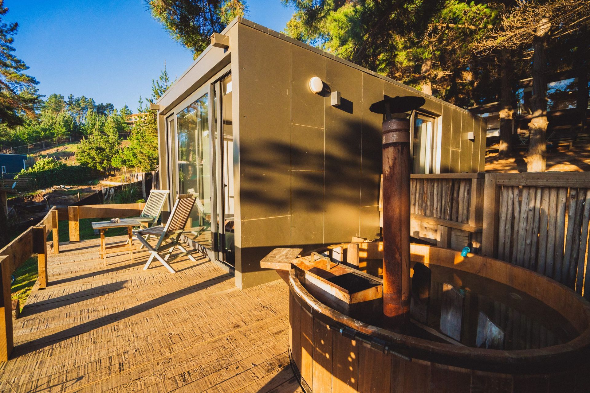 Un jacuzzi de leña se encuentra en una terraza de madera frente a una cabaña moderna rodeada de árboles al atardecer.