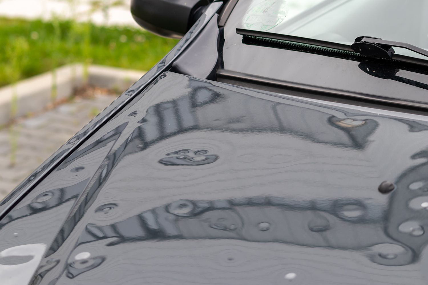 Close-up of a car hood with water droplets reflecting nearby buildings.