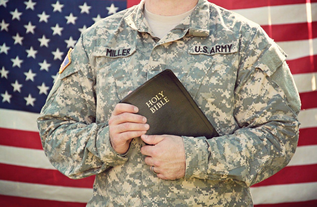 US Army soldier in camouflage holding a Bible in front of a U.S. flag