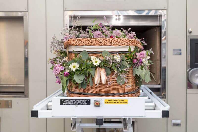 A flower-filled casket on a hospital morgue tray, surrounded by pink, white, and purple blooms