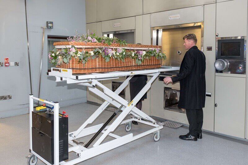 Man in a black coat pushing a scissor lift carrying a flower-filled coffin in a crematorium room