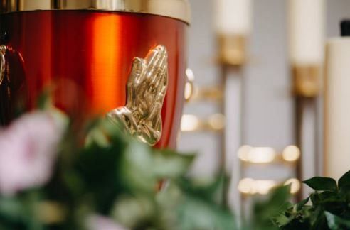 Red memorial candle with gold praying hands beside blurred flowers and candles