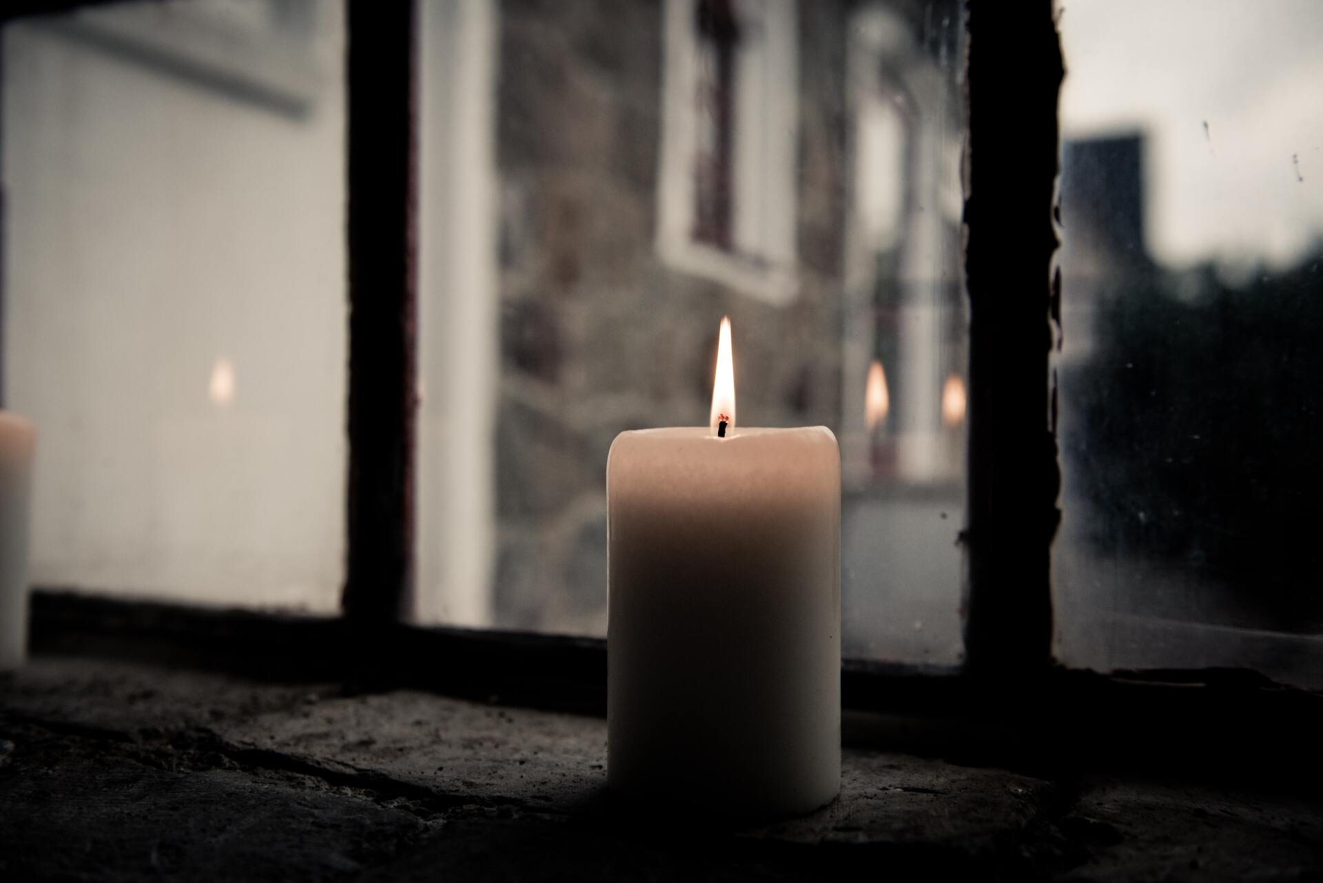 Lit white candle on a windowsill beside a rainy window, with a blurred building outside