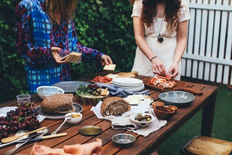 People serving food at an outdoor table laden with dishes, bread, and grapes