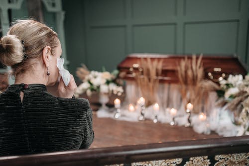 Person in black stands beside an open casket surrounded by white flowers and candles