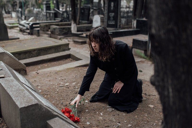 Person kneels by a grave, placing red flowers in a cemetery.