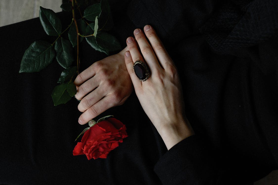 Hands holding a red rose against a dark background, with a black ring on one finger