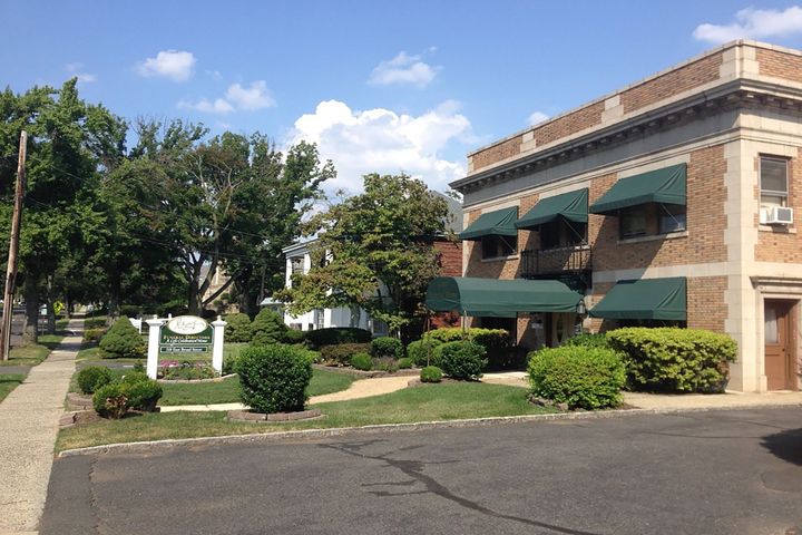 Brick office building with green awnings, trees, shrubs, and a paved parking lot under a blue sky