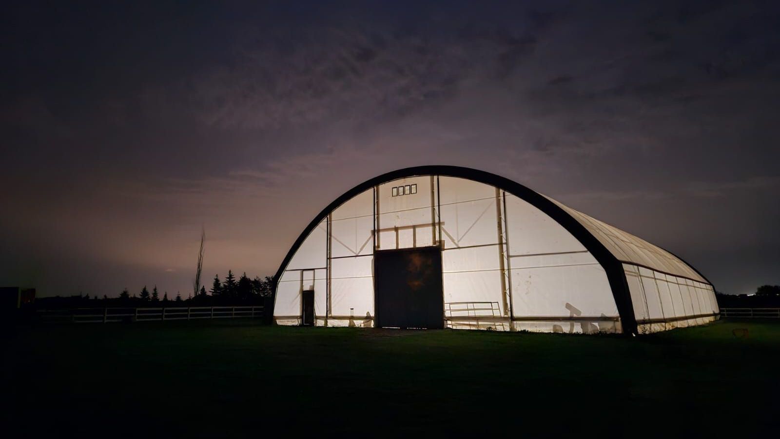 An illuminated, arched-roof building at dusk, in a field with a dark sky.