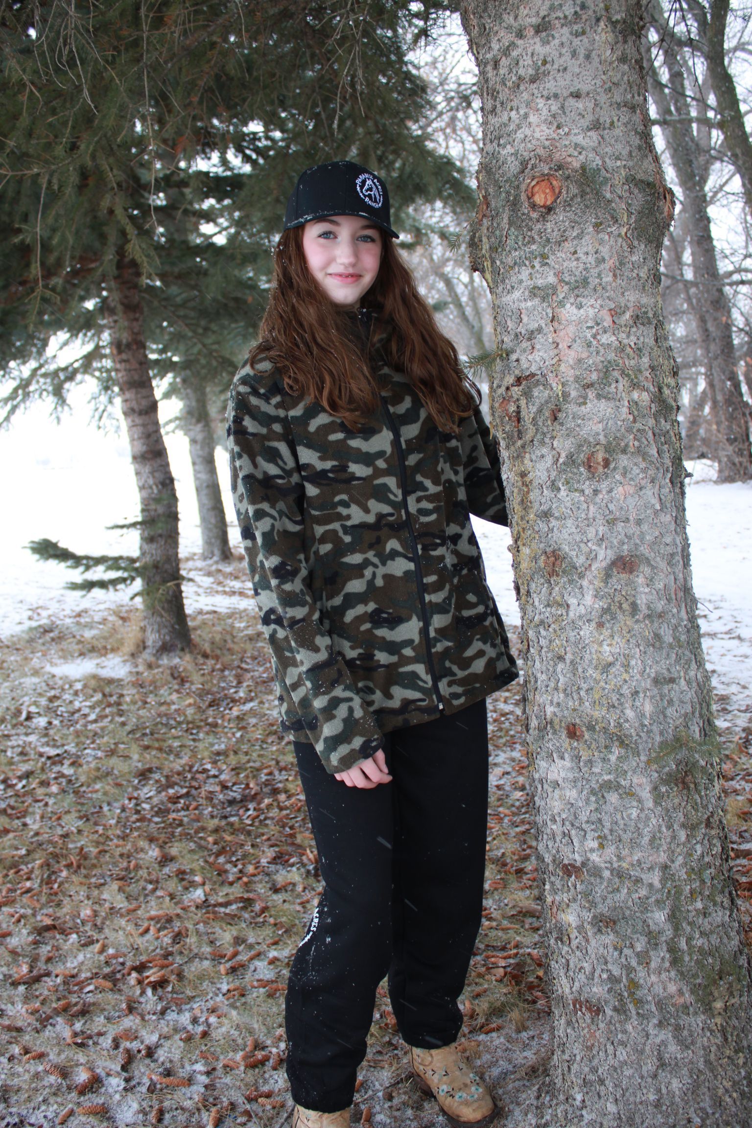 Smiling person in a black cap beside a tree outdoors in winter