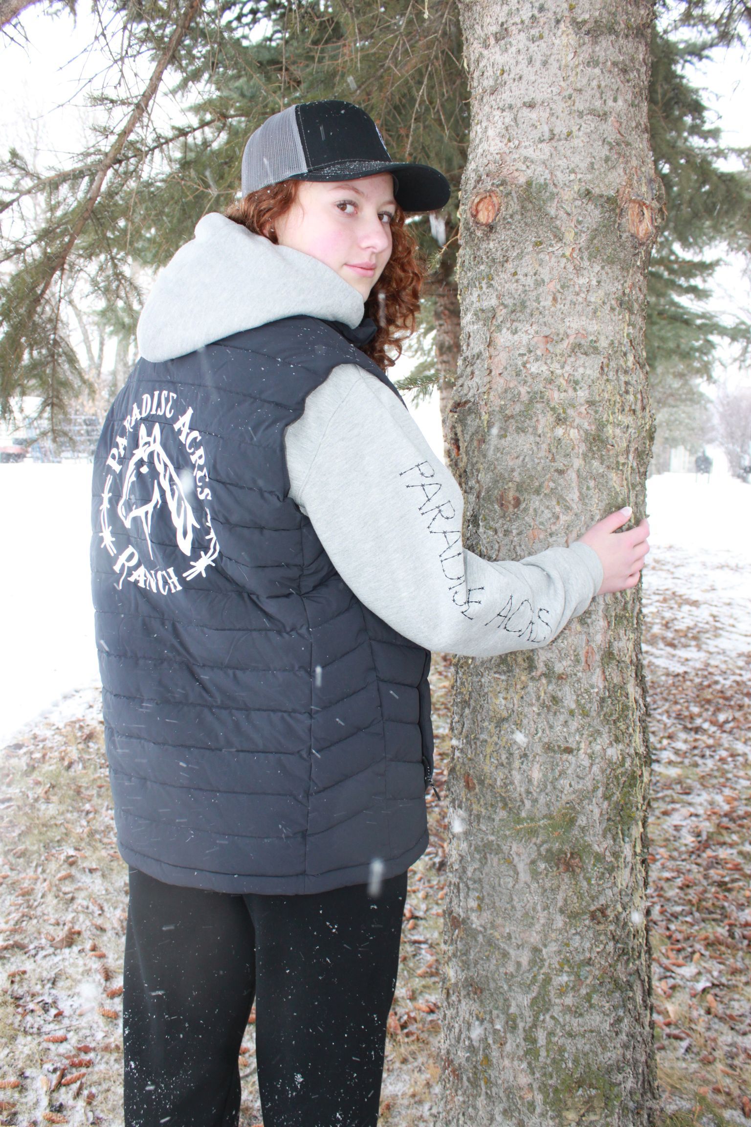 Person with curly red hair in winter clothes stands by a tree in the snow.