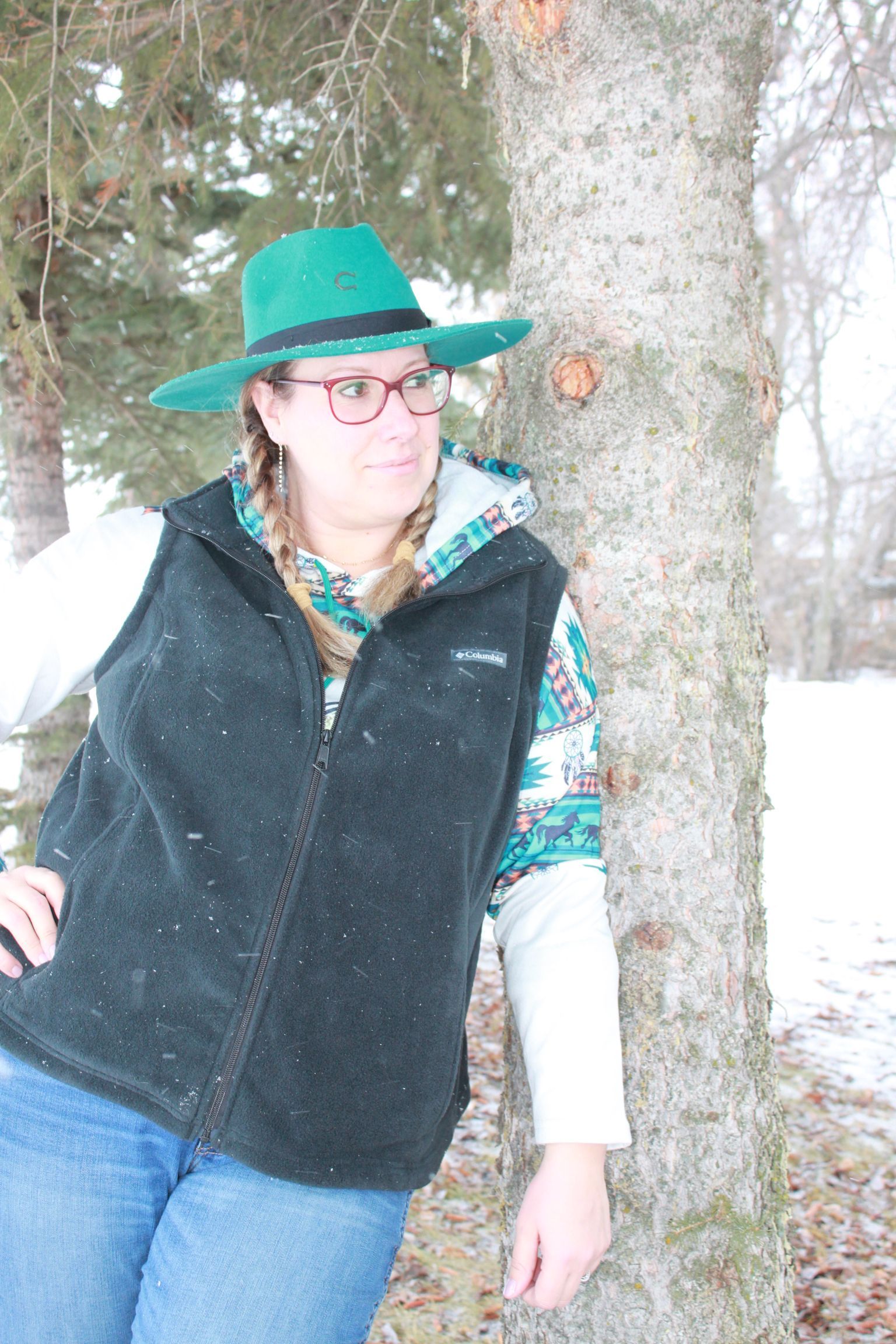 Woman in teal hat and vest with horse logo touches a tree in snowy field.