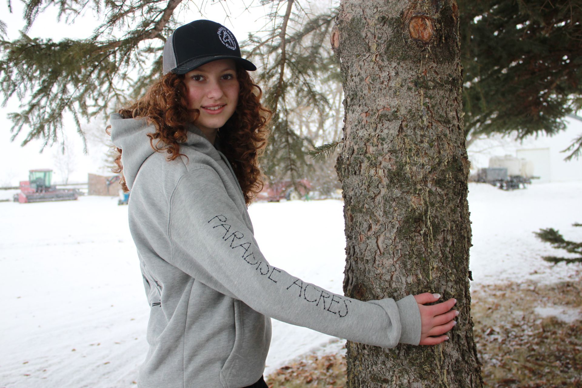 A person with curly red hair wearing a grey sweatshirt and black cap leans against a tree in a snowy outdoor setting.