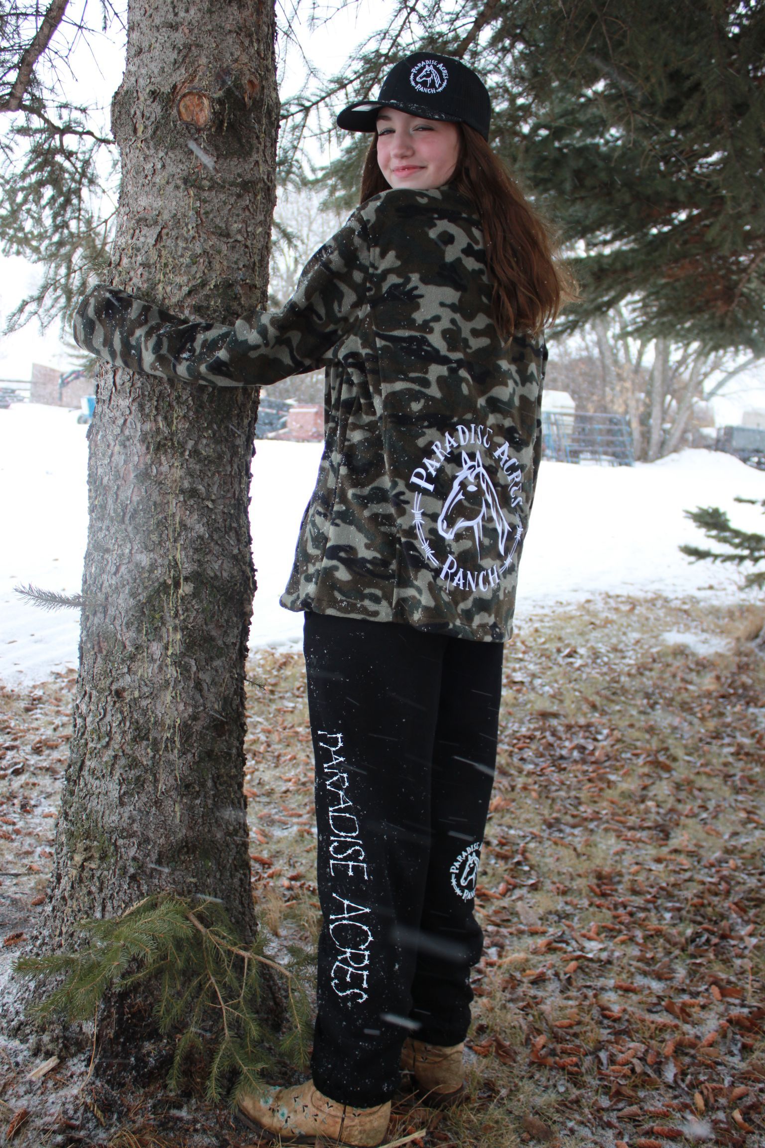 Teen girl in camo jacket, baseball cap, smiles next to a tree in a snowy outdoor setting.