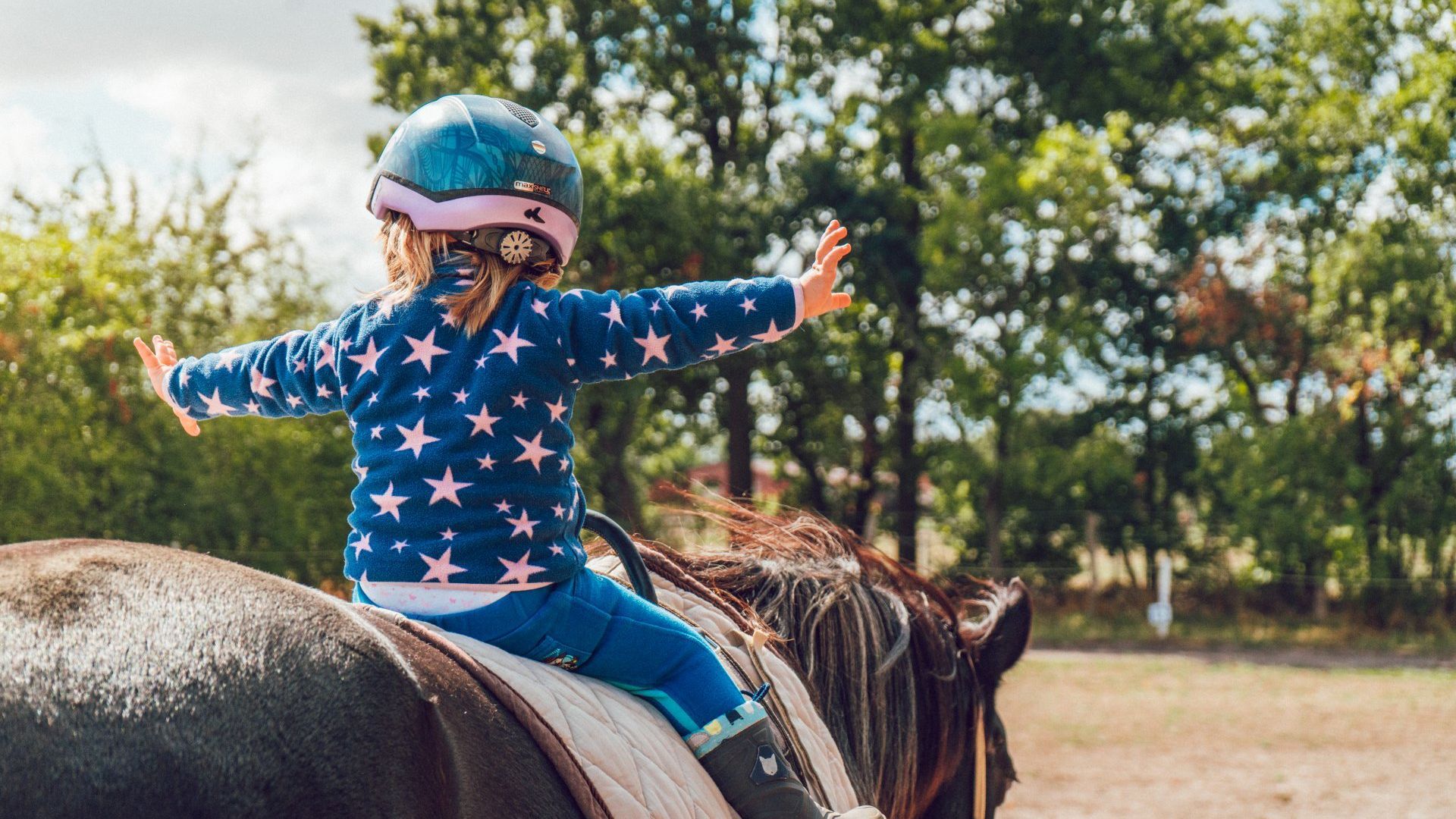 Girl with arms out, wearing a helmet, riding a horse in an outdoor setting.