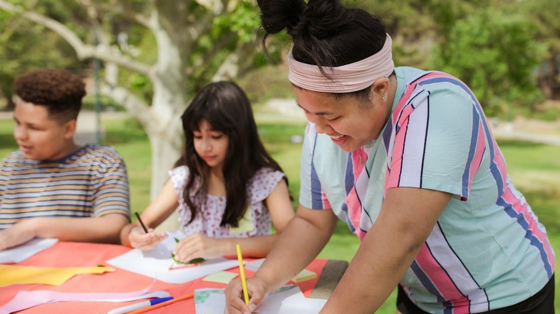 A group of kids drawing at a picnic table under a tree. A woman guides their work, smiling.